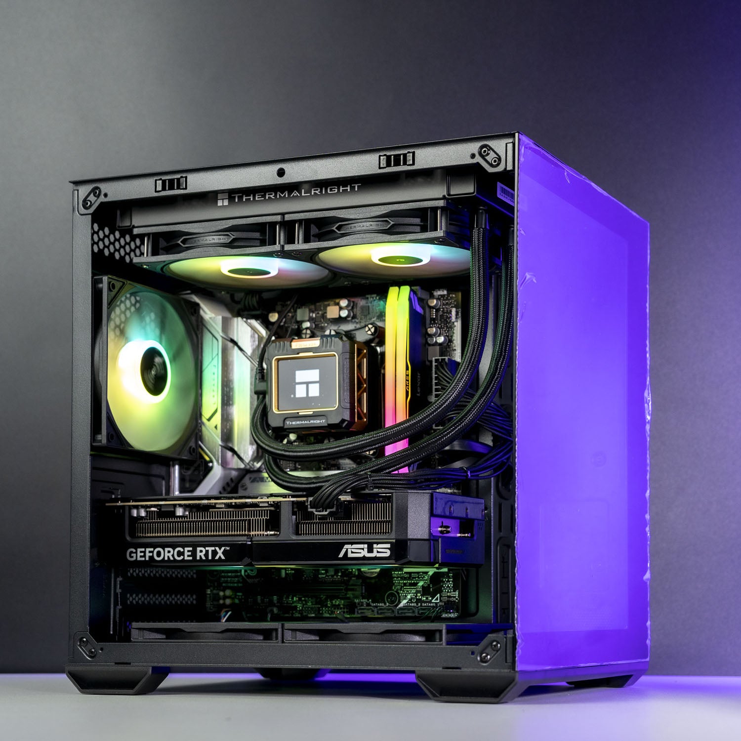 A high-end custom PC build in a black Thermaltake case with a clear side panel, showcasing internal components including an ASUS GeForce RTX graphics card, a liquid cooler with RGB lighting, and dual CPU coolers, set against a dark gray studio background.