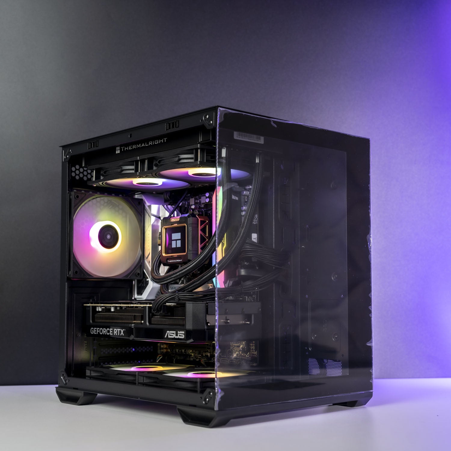 A high-end custom PC build in a black Thermaltake case with a clear side panel, showcasing internal components including an ASUS GeForce RTX graphics card, a liquid cooler with RGB lighting, and dual CPU coolers, set against a dark gray studio background.