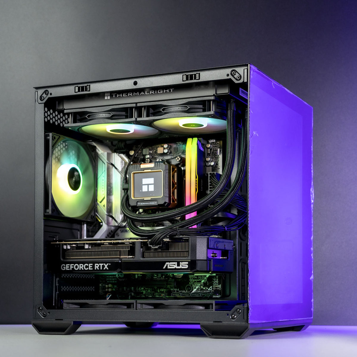 A high-end custom PC build in a black Thermaltake case with a clear side panel, showcasing internal components including an ASUS GeForce RTX graphics card, a liquid cooler with RGB lighting, and dual CPU coolers, set against a dark gray studio background.