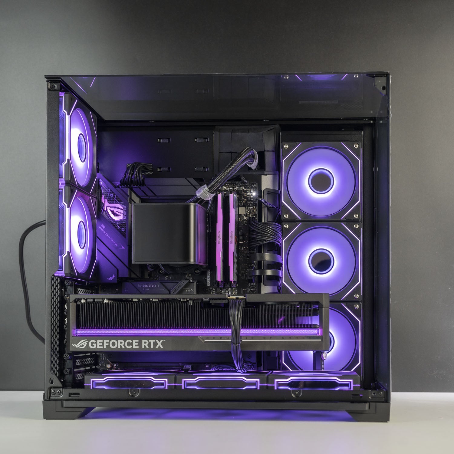 Open black computer case showing a custom PC build with a GeForce RTX graphics card, dual CPU coolers, and multiple RGB fans glowing with purple LED light against a dark gray background.